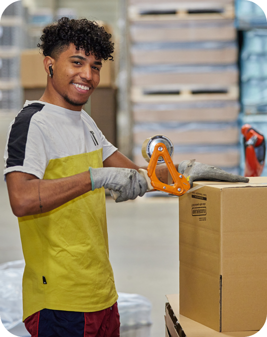 Mitarbeiter Palettenbau Lächelnder Mann in weiß-gelbem T-Shirt verschließt einen Versandkarton mit Klebeband