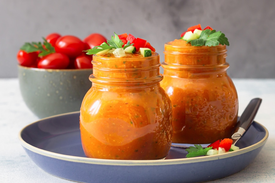 Rezept Gazpacho Stimungsbild: Zwei Schraubverschlussgläser gefüllt mit roter Gazpacho arrangiert auf einem blauen Teller, im Hintergrund eine Schale mit Tomaten